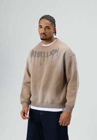 Beige sweatshirt met "REBELLION" gedrukt in een druppelend lettertype. Voorzien van een ronde hals, geribbelde manchetten en een gestructureerde afwerking. Gecombineerd met een wit overhemd.