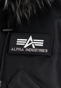 Crna jakna od tkanine s umjetnim krznom na ovratniku. Ima izvezeni crni i bijeli pečat s natpisom "ALPHA INDUSTRIES" i stiliziranim logotipom.