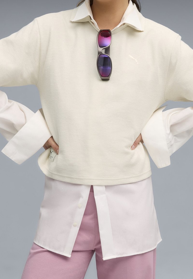 Personne portant une chemise blanche superposée et un pull avec des lunettes de soleil violettes accrochées au pull, les mains sur les hanches, pantalon rose visible en dessous.