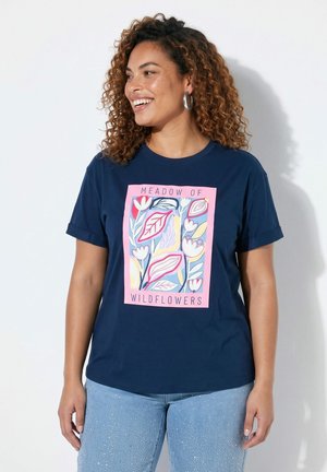 Femme aux cheveux bouclés souriante, portant un t-shirt bleu avec un motif floral rose et le texte "Meadow of Wildflowers", ainsi qu'un jean bleu clair avec des détails scintillants.