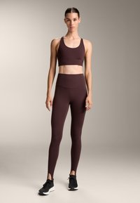 Un sutien sport maro și colanți cu talie înaltă, cu slituri laterale. Materialul este elastic și neted, asortat cu încălțăminte sport neagră.