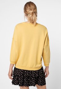 Sudadera amarilla de tres cuartos, de corte relajado y con dobladillo acanalado, combinada con una falda negra con volantes que presenta pequeños patrones florales en rosa.