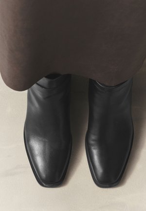 Zwarte leren enkellaarzen met vierkante tenen gedragen onder een bruine rok op een lichte vloer.