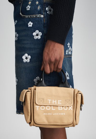 Beige Canvas-Handtasche mit schwarzen Lederakzenten, versehen mit dem Aufdruck "DIE WERKZEUGKISTE" in Weiß und mehreren Taschen zur Aufbewahrung.