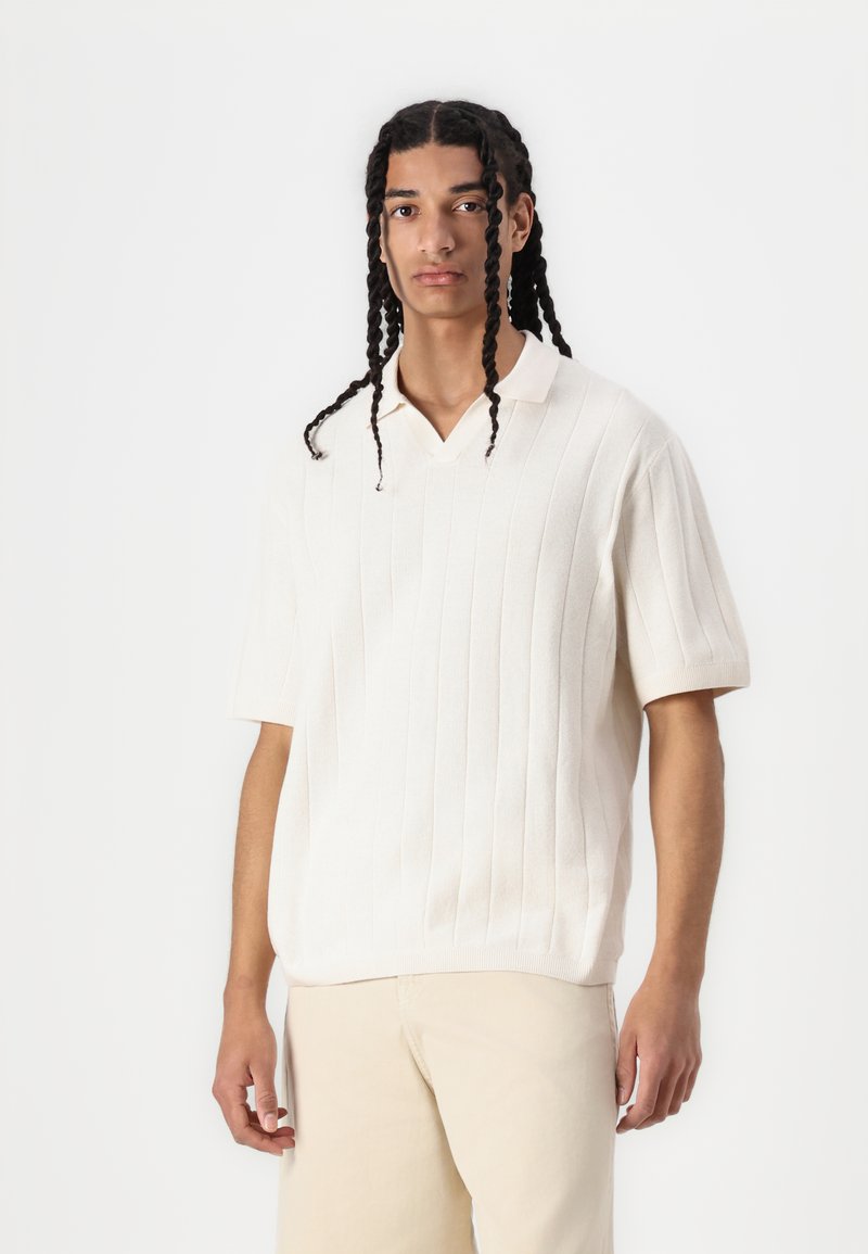 Kortermet, kremfarget polo-skjorte med ribbet tekstur og V-hals design. Brukt med lysebeige bukser.
