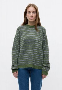 Young woman with long light brown hair wears green and gray striped sweater and blue jeans, standing against a plain white background.