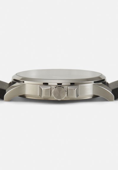 Montre en acier inoxydable avec un boîtier rond argenté, une lunette lisse, une couronne texturée et un bracelet en cuir noir. Le verre reflète légèrement la lumière.