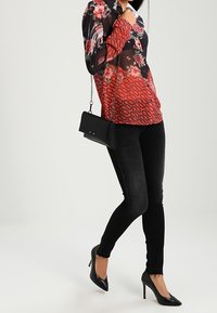 Blusa floreale rossa e nera fluida con tessuto trasparente, abbinata a jeans skinny neri e tacchi a punta neri. Piccola borsa a tracolla nera.