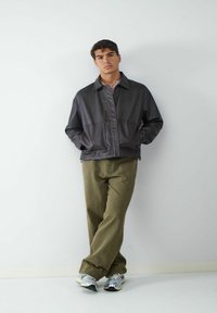 Donkerleren jas met een ritssluiting aan de voorkant, puntige kraag en twee achterzakken, gecombineerd met lichtolijfgroene broek en metallic sneakers.