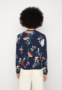 Personne aux cheveux bouclés portant un haut à manches longues bleu marine à motifs floraux et un pantalon beige clair, debout de dos face au spectateur.