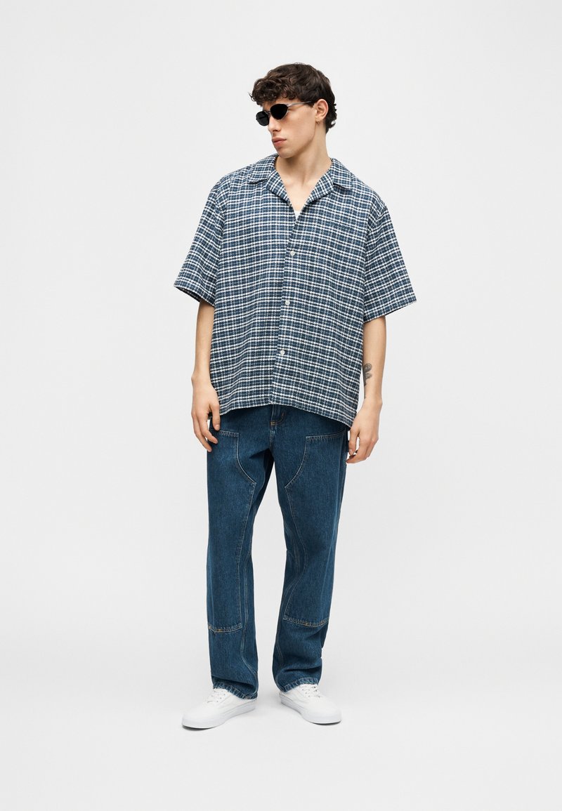 Joven con gafas de sol negras, camisa de manga corta a cuadros azul y blanco, vaqueros azules y zapatillas deportivas blancas, de pie frente a un fondo liso.