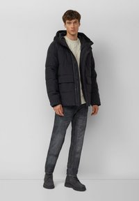 Schwarze gesteppte Jacke mit Kapuze, ausgestattet mit zwei Vorder Taschen und Reißverschluss. Das Modell trägt einen leicht gestrickten Pullover und dunkle Jeans.