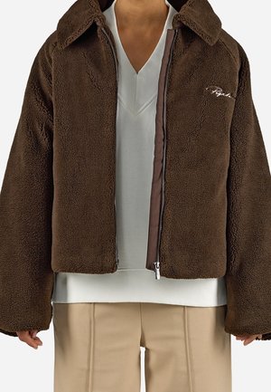 Osoba ubrana w brązową polarową kurtkę na zamek na białym swetrze z dekoltem w serek oraz beżowe spodnie, ukazana od szyi do bioder.