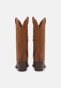 Bottes de cow-boy marron à bout pointu, avec des coutures décoratives et des languettes pour les enfiler. Elles présentent un talon empilé et une surface en cuir lisse.