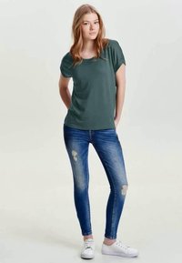 T-shirt de mangas curtas verde-escuro combinada com jeans skinny azul desbotado com detalhes desgastados, complementada por ténis brancos.