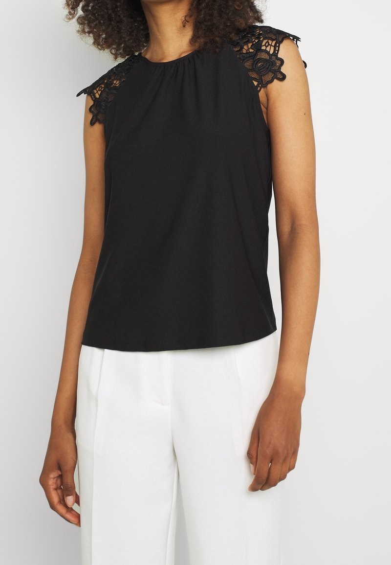 Haut noir sans manches avec un col froncé et des détails en dentelle sur les épaules. Tissu doux ; coupe décontractée ; sans attaches visibles.