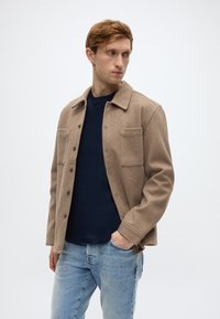 Πουκάμισο-μπολερό σε ταμπά χρώμα από μείγμα μαλλιού με κούμπωμα με κουμπιά στο μπροστινό μέρος, δύο τσέπες στο στήθος και κανονική εφαρμογή, σε συνδυασμό με μπλε κοντομάνικο μπλουζάκι λαιμόκοψης και μπλε τζιν.