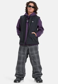 Gilet nero senza maniche sopra un felpa viola, abbinato a pantaloni baggy grigi a quadri e scarpe sportive nere. Gli accessori includono occhiali da sole scuri.