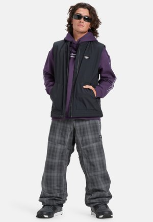 Gilet sans manches noir sur un sweat à capuche violet, associé à un pantalon baggy à carreaux gris et des chaussures de sport noires. Les accessoires comprennent des lunettes de soleil sombres.