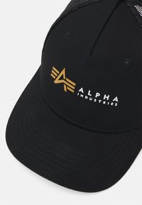 Fekete sapka ívelt karamellyel, az elején arany logóval és fehér "ALPHA INDUSTRIES" felirattal. Texturált hálós hátsó panel.