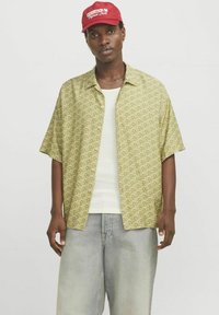 Kurzarm-Hemd mit Knöpfen in Hellgrün mit geometrischen Mustern; getragen über einem weißen, strukturierten Tanktop und kombiniert mit hellen Jeansshorts.