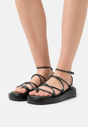 Černé platformové sandály se zeleným proužkovým designem, vyznačující se tenkými šňůrkami a nastavovacími pásky kolem kotníku. Hladký povrch s otevřenou špičkou.