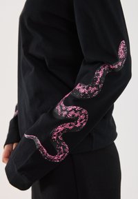 Chemise noire à manches longues avec un graphisme de serpent rose sur la manche, fabriquée en tissu coton doux avec une texture lisse.