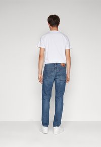 Camiseta blanca de hombre y jeans de mezclilla azul. Los jeans tienen un corte clásico, cinco bolsillos y un parche en la cintura trasera. Zapatillas blancas completan el conjunto.