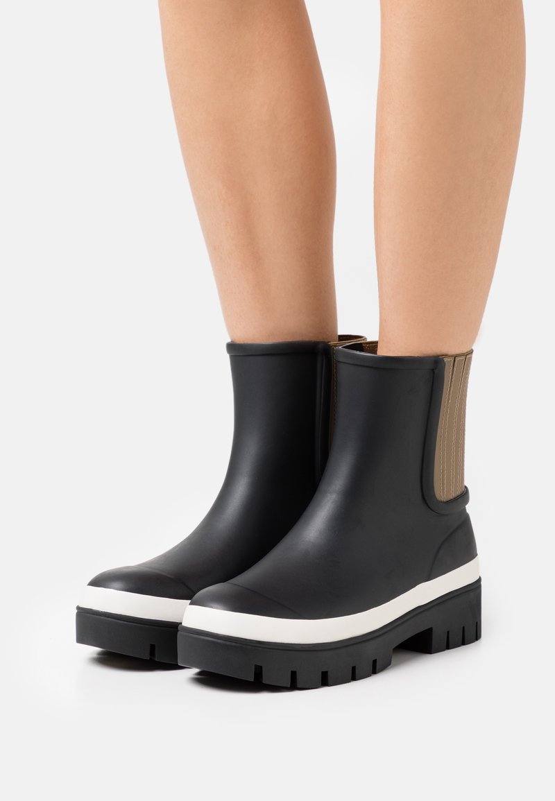 Černé gumové kotníkov é boty s texturovanou béžovou elastickou stranou, bílou mezipodešví a robustní černou podešví. Hladký povrch s kulatou špičkou.