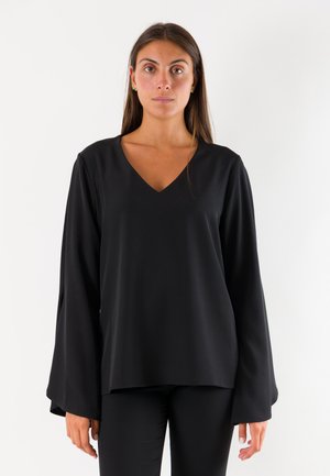 Donna con lunghi capelli castani che indossa una maglia nera larga a maniche lunghe con scollo a V e pantaloni neri, in piedi davanti a uno sfondo bianco semplice.