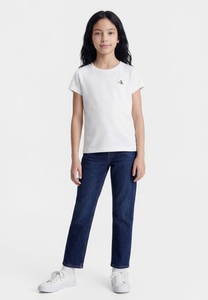 Jeune fille debout, portant un t-shirt blanc Calvin Klein, un jean bleu foncé, des chaussettes blanches et des baskets blanches sur un fond uni.