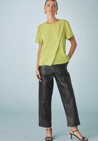 Blusa verde lime a maniche corte con un design drappeggiato abbinata a pantaloni larghi in pelle nera a vita alta e sandali neri con cinturini.