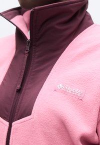 Κοντινό πλάνο ενός ροζ και βυσσινί φλις μπουφάν Columbia Sportswear με φερμουάρ, με μπάλωμα λογότυπου στο στήθος.