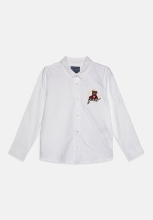 Camicia bianca a maniche lunghe con colletto classico, chiusura con bottoni e design di un orso ricamato con il testo "Guess" sul petto sinistro.