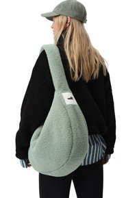 Zielona torba typu hobo z pluszowego materiału o miękkiej teksturze, w oversize'owym kształcie, z regulowanym paskiem, z małą białą metką z przodu.