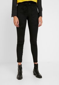 Svarta, figurnära byxor i ett mjukt, strukturerat material med framfickor med dragkedja och hög midja. Bärs med ankelstövlar, vilket framhäver benen.