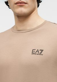 Εγγύς λήψη ενός ατόμου που φοράει μπεζ μπλουζάκι με μικρό μαύρο λογότυπο "EA7 Emporio Armani" στο αριστερό στήθος.