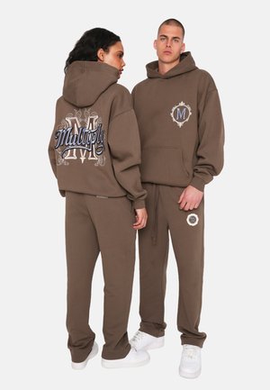 Bruine oversized hoodies en sweatpants hebben geborduurd "Multiply"-tekst op de achterkant, met accenten en logo-details in contrasterende kleuren.
