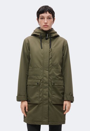 Person iført en lang olivengrøn hætteparka med sorte snore og store forlommer, stående mod en ensartet lys baggrund.