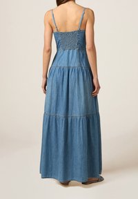 Robe maxi en denim avec des bretelles fines, un corsage smocké et une jupe à volants fluide dans un bleu moyen. Des sandales plates complètent le look.