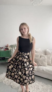 Frau mit langen blonden Haaren, die ein ärmelloses schwarzes Oberteil und einen schwarzen Rock mit beigen Blumenmustern trägt, steht in einem hellen Wohnzimmer.