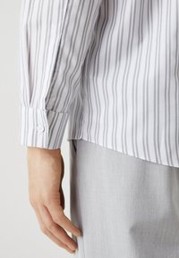 White long-sleeve shirt with thin gray vertical stripes, featuring a buttoned cuff and a straight hemline, paired with light gray trousers.
