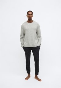 Ανοιχτό γκρι, πουλόβερ με μίξη μαλλιού και στρογγυλή λαιμόκοψη, σε συνδυασμό με στενά, μαύρα βαμβακερά παντελόνια. Το μοντέλο είναι ξυπόλητο και στέκεται όρθιο.