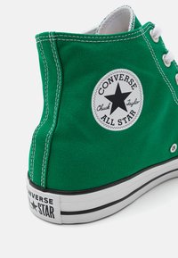 Roheline canvas kõrge varbaga spordijalanõud valgete nööpide, musta ja valge tähe logoga, õmmeldud detailidega ja valge kummeeritud tallaga, millel on must riba.