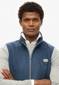 Granatowa kamizelka polarowa z zamkiem z przodu, szara bluzka z długim rękawem pod spodem oraz naszywka z logo na lewej piersi. Miękka struktura i luźny krój.