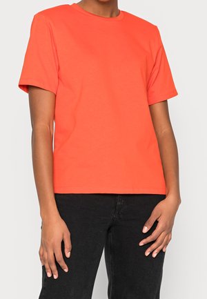 Person wearing a plain bright orange short-sleeve t-shirt and black pants, standing against a plain light background.
