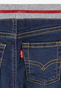 Jean en denim bleu avec des coutures jaunes et une ceinture élastique grise ornée d'une bande rouge, montrant des poches arrière et une étiquette rouge sur une poche.