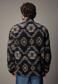 Veste en polaire texturée avec un motif géométrique noir, beige et bleu, dotée d'un col haut et d'une coupe décontractée.