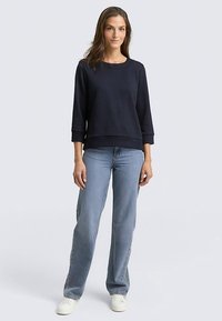 Marineblauwe sweatshirt met textuur en driekwart mouwen, gecombineerd met lichtblauwe jeans met rechte pijpen en witte sneakers.