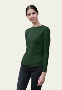 Pull vert à manches longues avec un col rond, design ajusté et texture lisse. Porté avec un jean noir. Fond dans une couleur neutre claire.